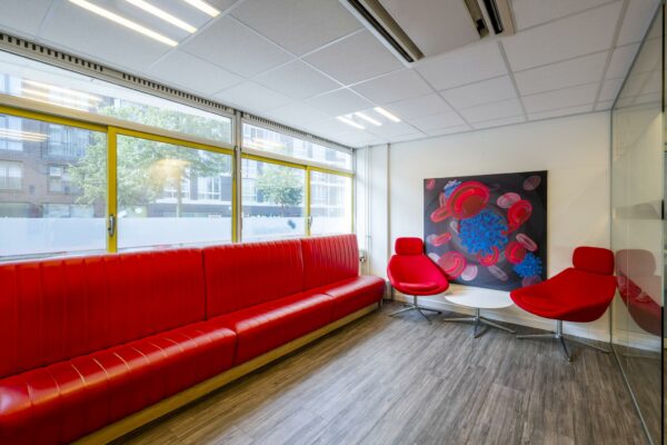 Wachtkamer met rood meubilair, grote ramen en een opvallend schilderij van rode bloedcellen en een virus aan de Jan Tooropstraat.